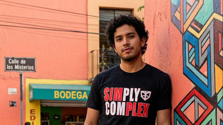 Person wearing a 'Simply Complex' t-shirt leaning against a colorful wall with a vintage car and storefronts in the background.